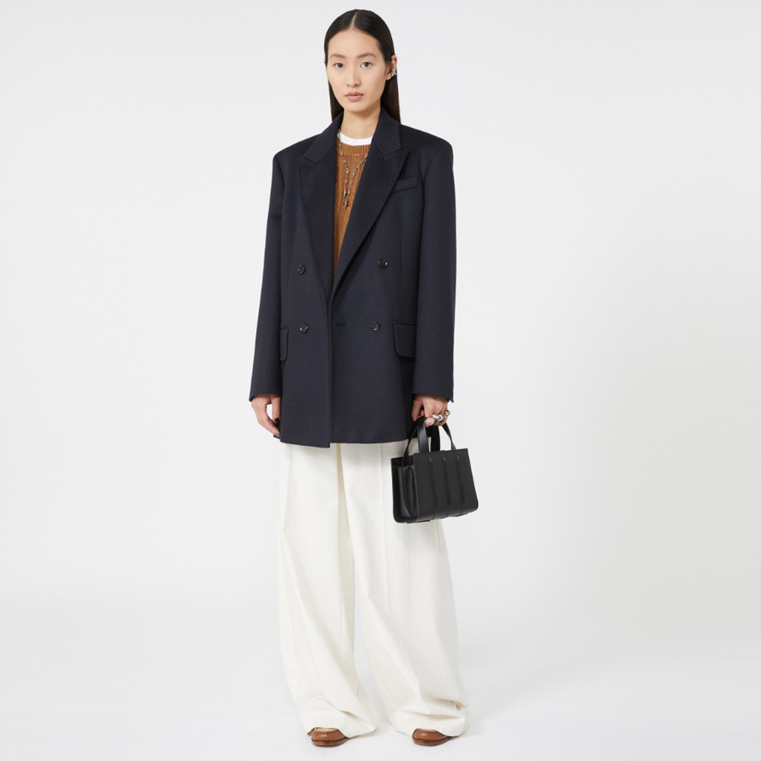 Model wearing a dark coat and white pants holding a black handbag on a white background