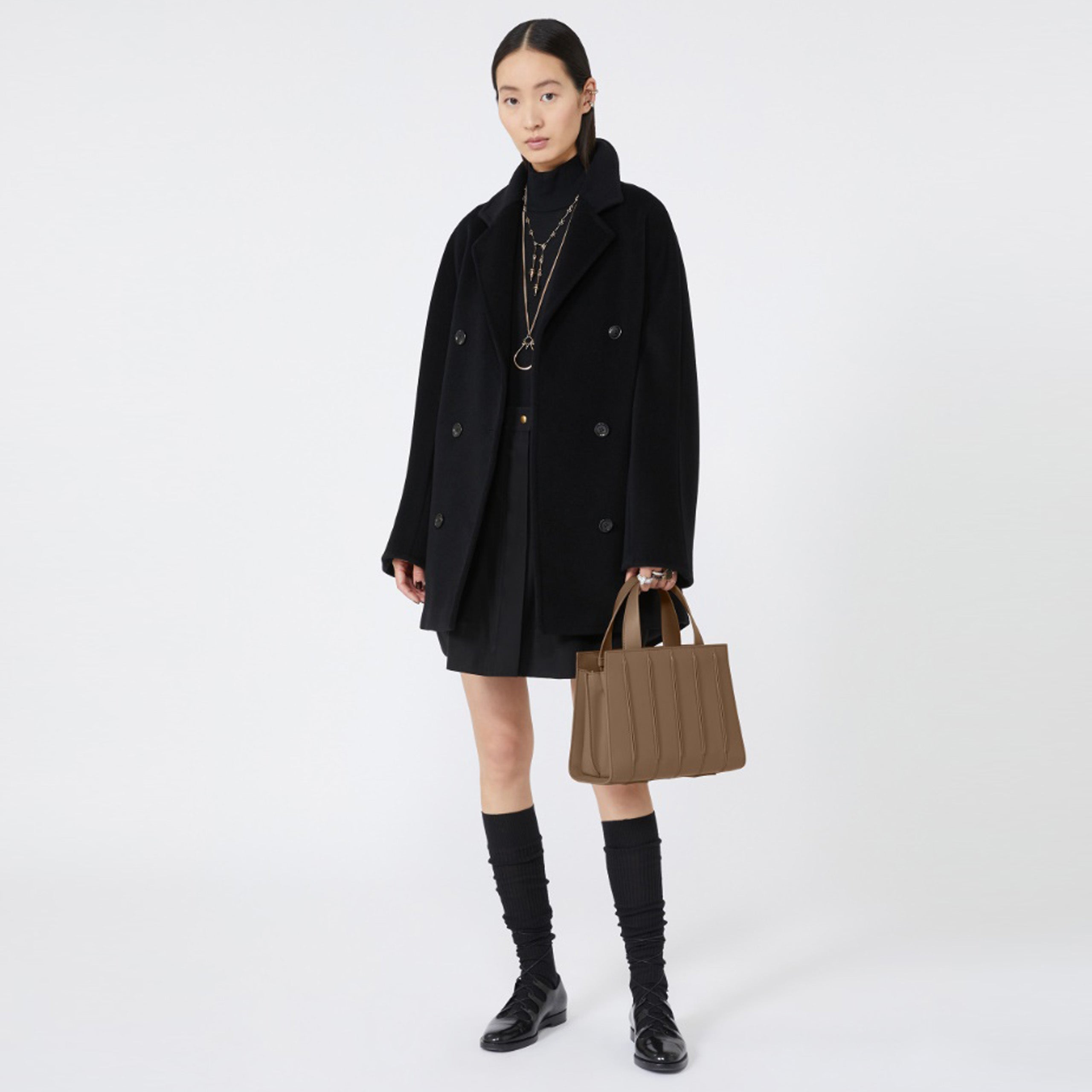 Model wearing a black coat and holding a camel bag on a white background