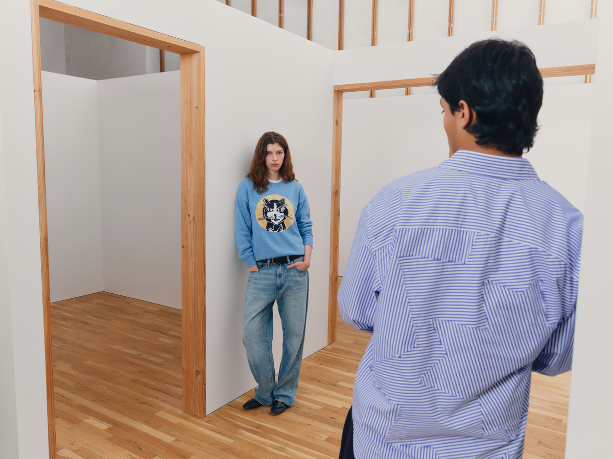 A model in a light blue crewneck sweatshirt with a graphic print inspired by Roy Lichtenstein's Laughing Cat and loose-fit jeans stands in a minimalist room with light wood floors and white walls, facing another model from behind wearing a blue and white striped button-down shirt inspired by Roy Lichtenstein's Standing Explosion