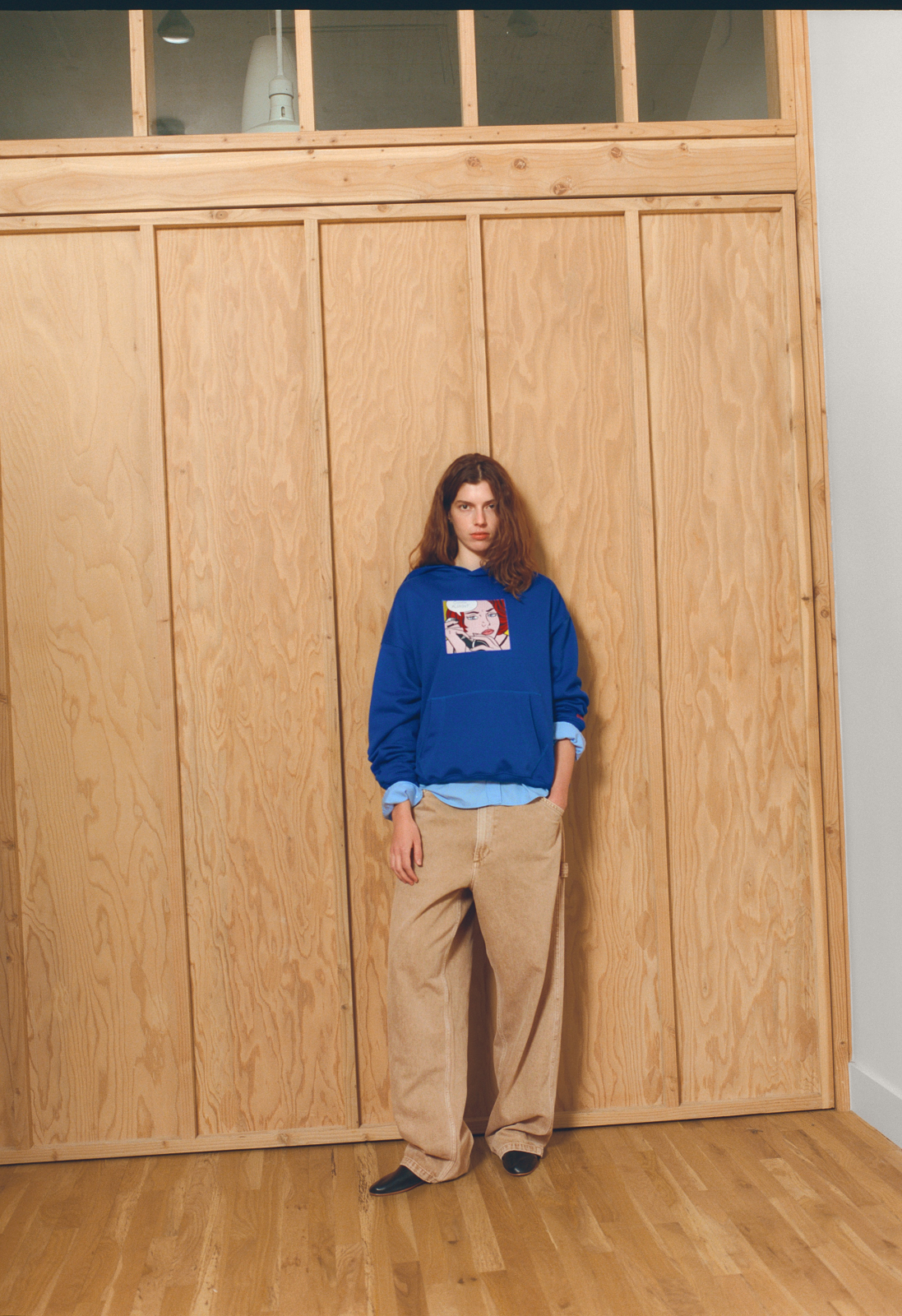 A model stands in front of a large wooden door wearing a blue sweatshirt with a graphic print inspired by Roy Lichtenstein's Ohhh... Alright... on the chest, a light blue shirt underneath, beige pants, and dark shoes
