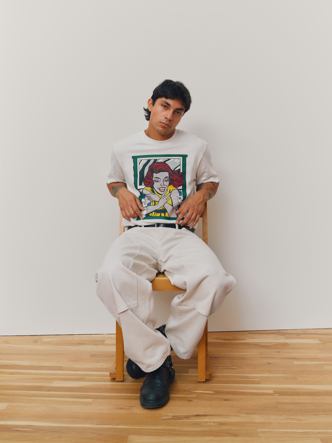 A model sits on a wooden chair against a plain off-white wall, wearing a white graphic t-shirt featuring Roy Lichtenstein's Girl in Window, loose white pants, and black boots