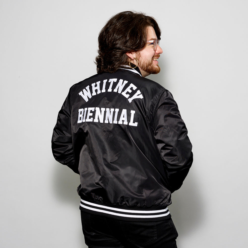 Model wearing 100% polyester black stadium jacket with "Whitney Biennial" in white text across the back