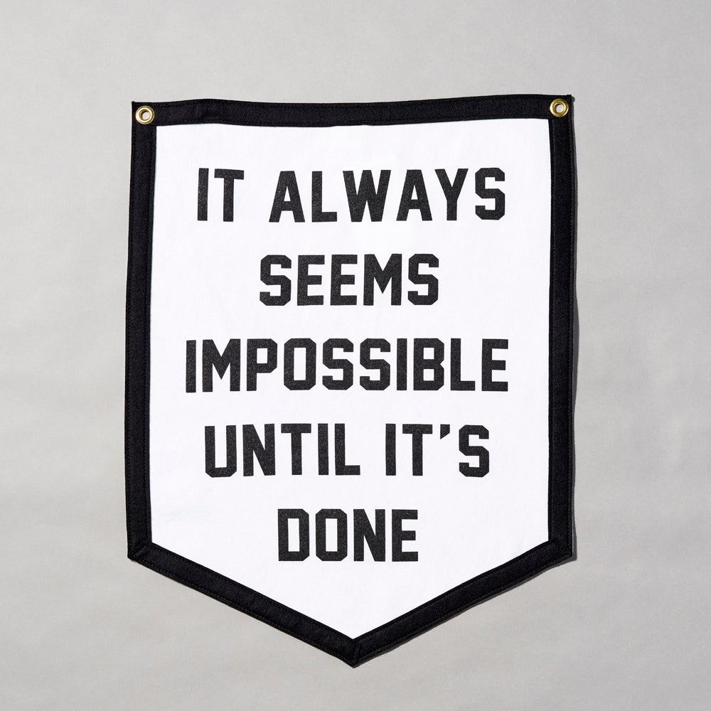 Screen printed felt flag with black border and white body. Text reads "It Always Seems Impossible Until It's Done" in black text. Measures 18" x 23