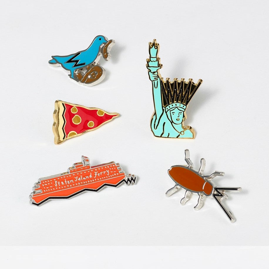 Assortment of hard enamel and metal pins illustrated by Tamara Shopsin, featuring iconic New York landmarks; Pigeon, Lady Liberty, Roach, Staten Island Ferry, and Pizza slice. 