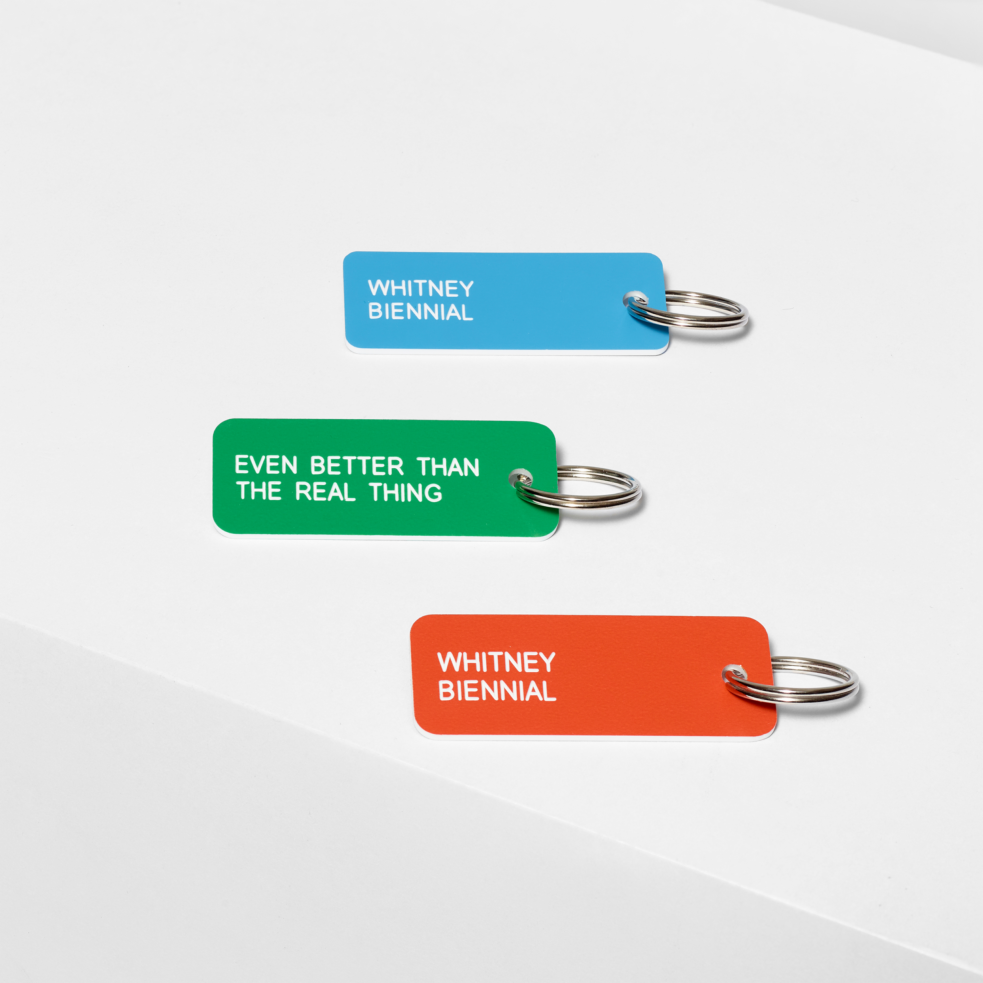 Assortment of etched acrylic keychains in various colors featuring the text Whitney Biennial on one side and Even Better Than The Real Thing on the other