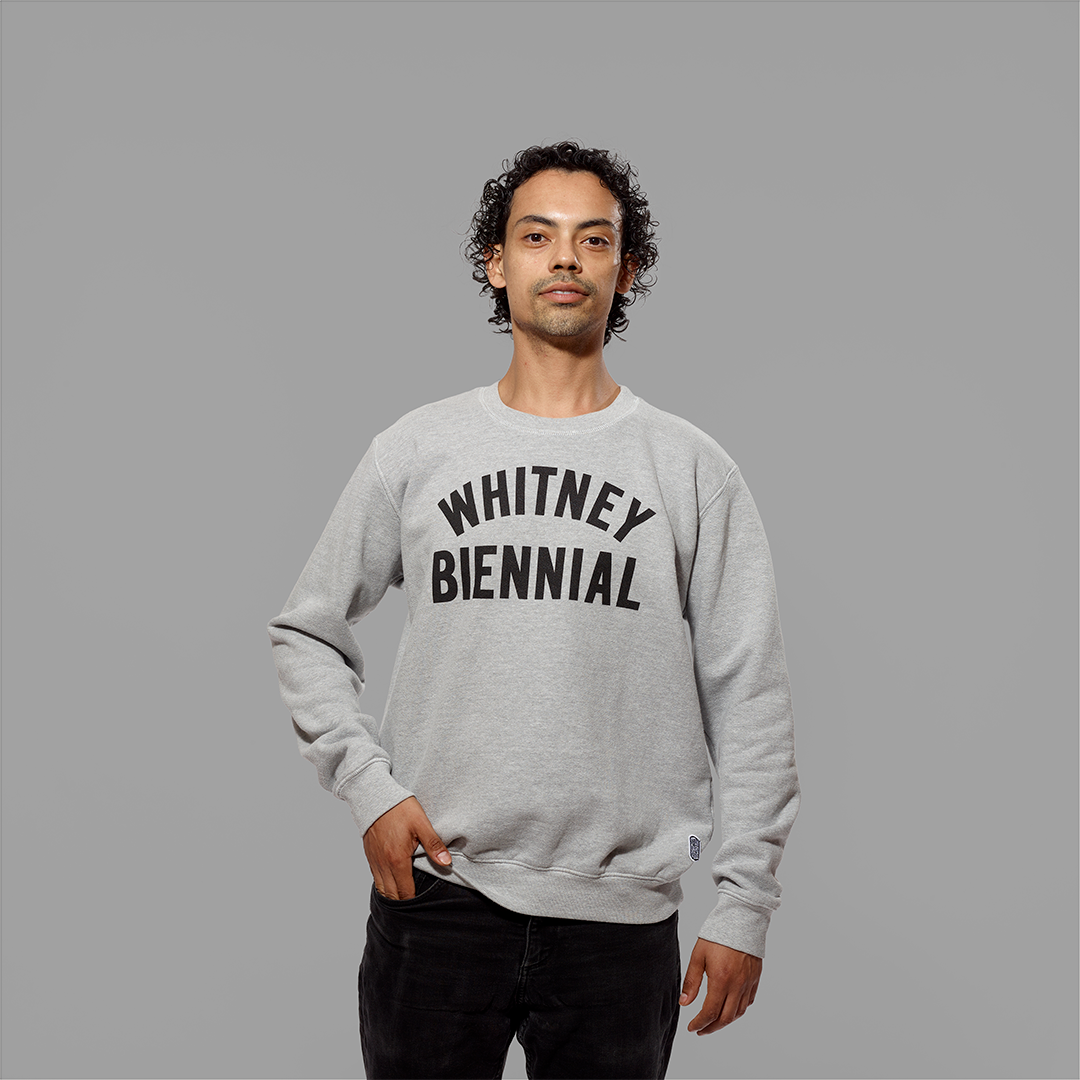 Model wearing a 100% cotton gray crew sweatshirt with Whitney Biennial in black text