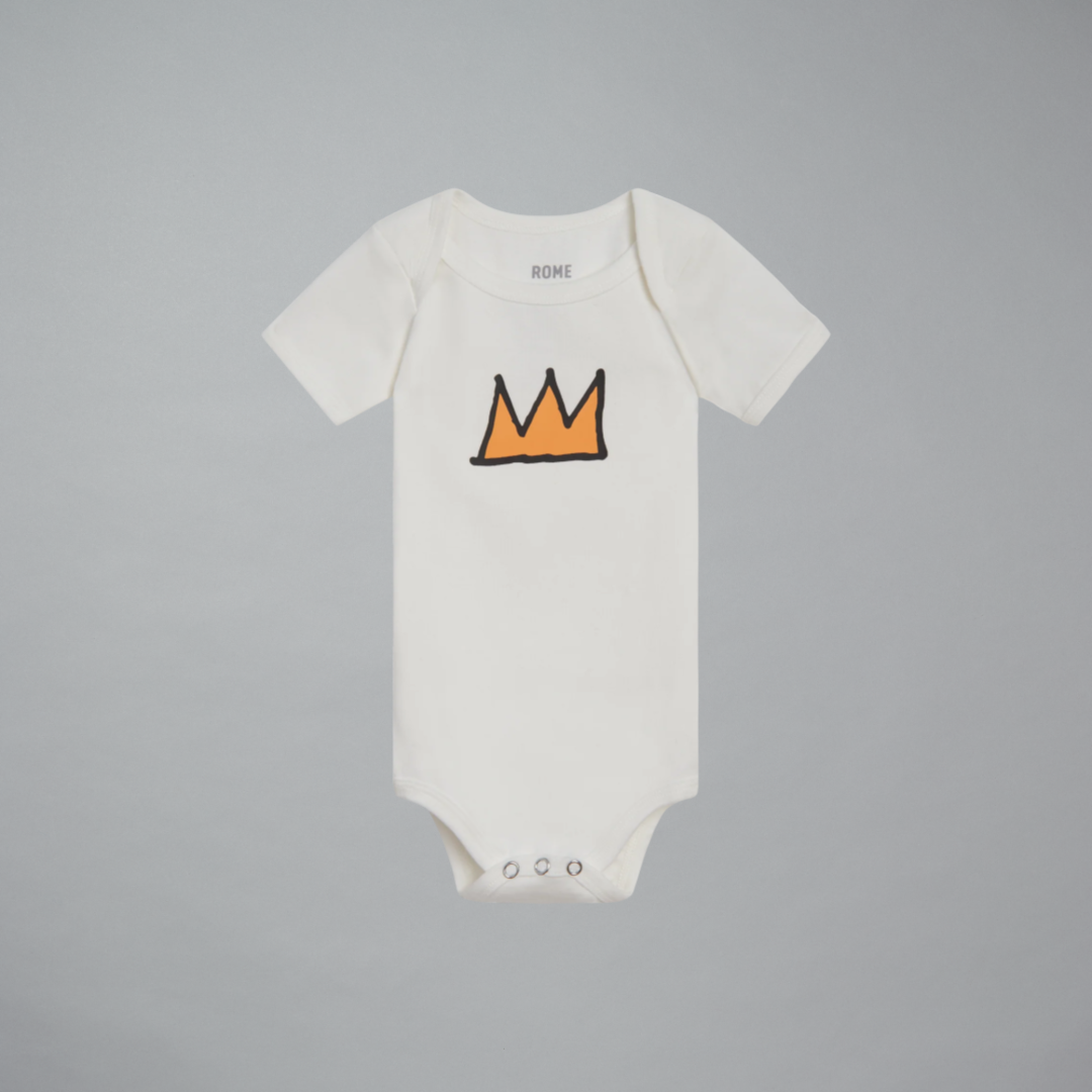 An off-white short-sleeve baby onesie with Basquiat's gold crown graphic outlined in black on the chest, displayed flat against a solid gray background.