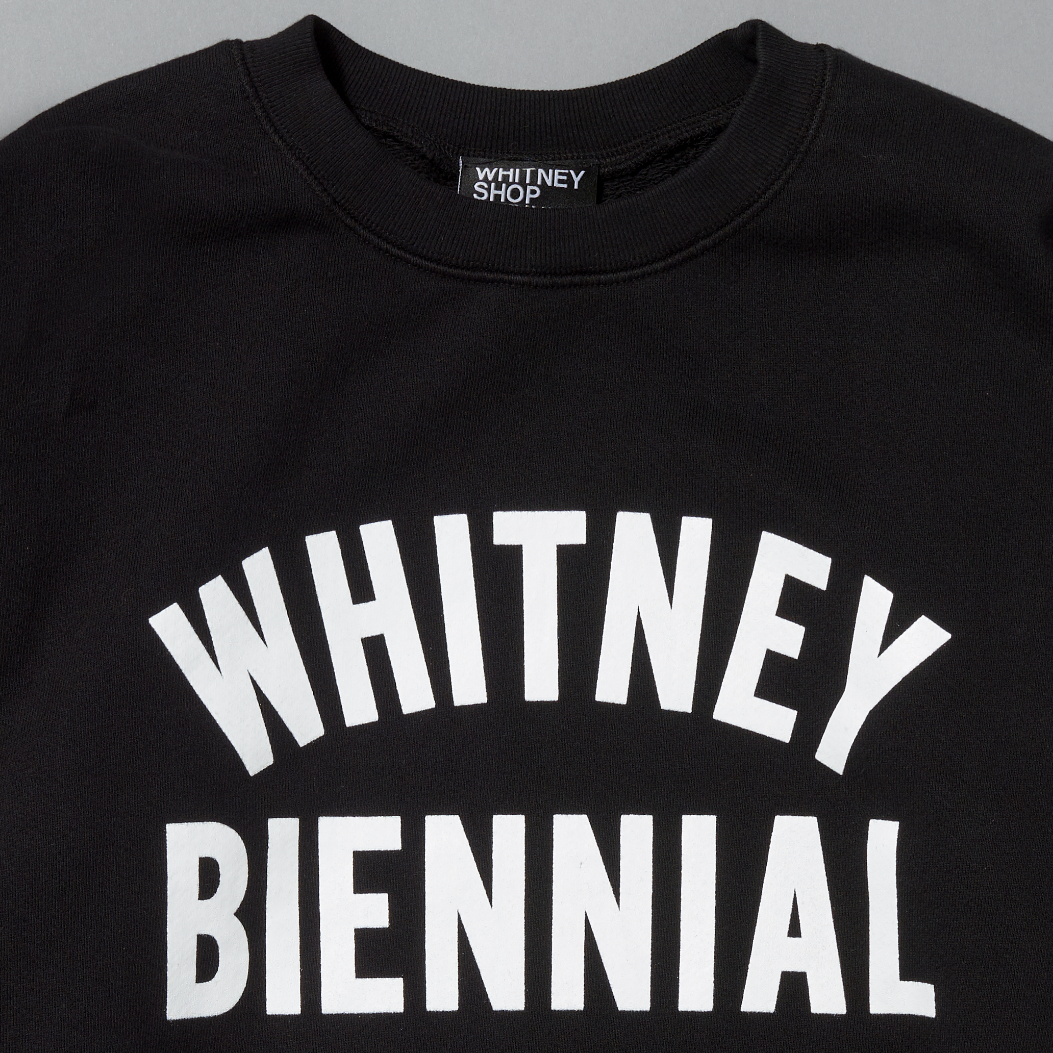 Black sweatshirt with 'WHITNEY BIENNIAL' white text on a gray background