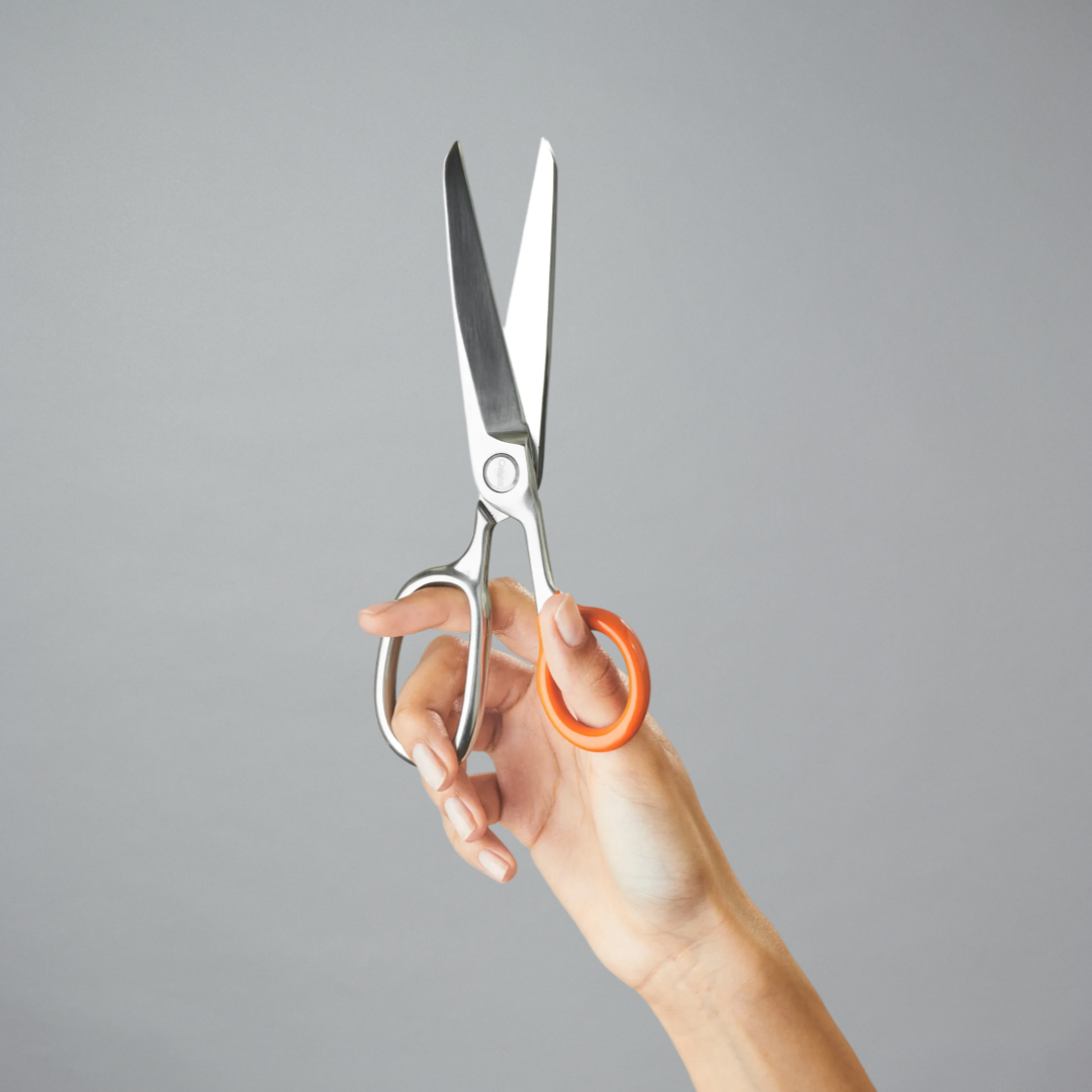 A hand holding a pair of large scissors with silver metal blades and bright orange handles against a gray background.