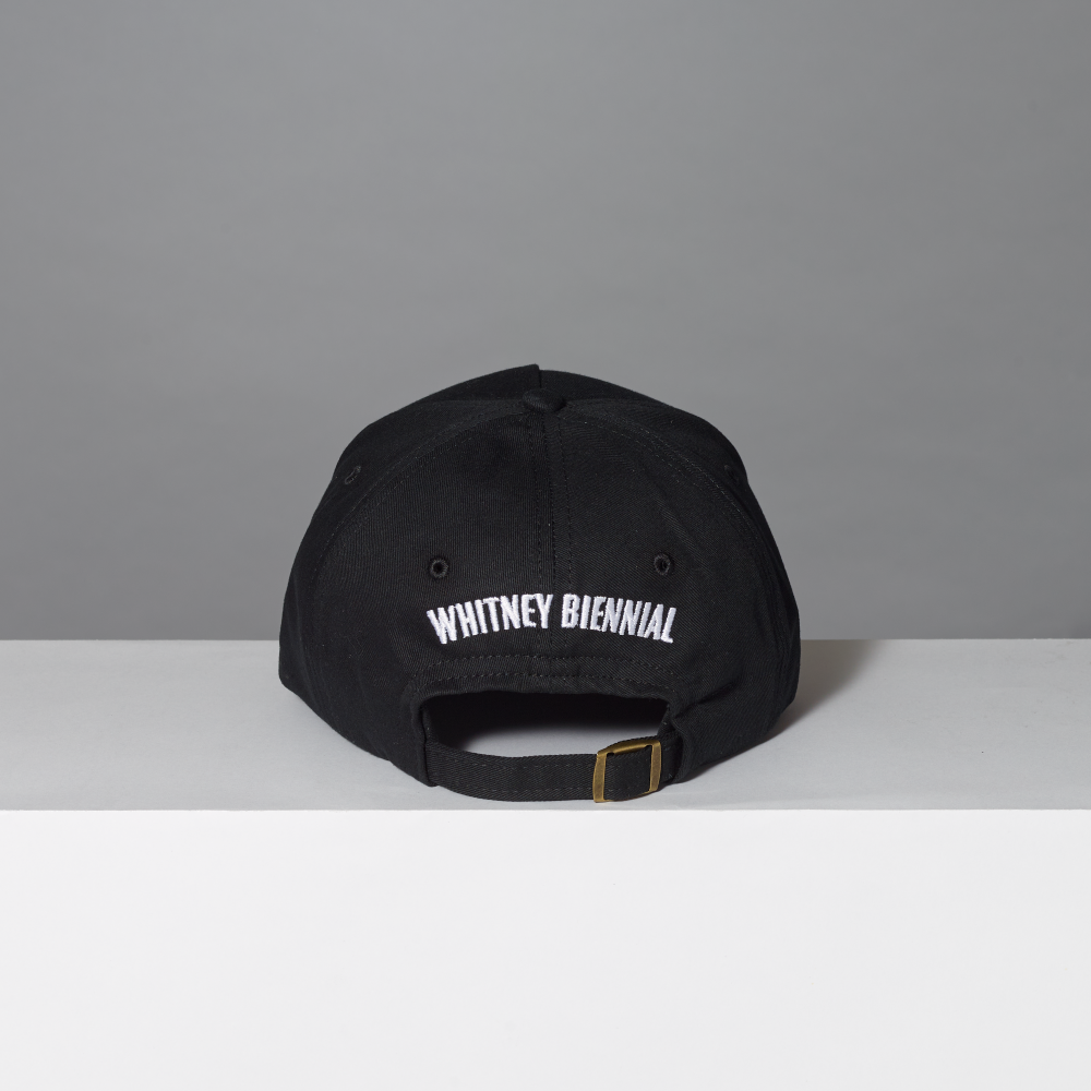 A black baseball cap with 'Whitney Biennial' embroidered in white block letters on the back, set against a neutral gray background.