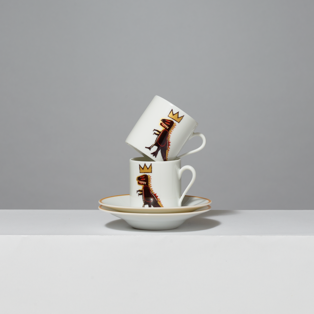 Two white ceramic espresso cups with matching saucers stacked together. The cups feature a printed illustration of Basquiat's Pez Dispenser dinosaur wearing a gold crown, set against a clean gray background.