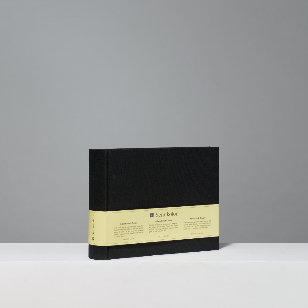 A black hardcover photo album standing upright on a white surface against a light gray background. A pale yellow paper band wraps around the middle, with the brand name 'Semikolon' printed on it.