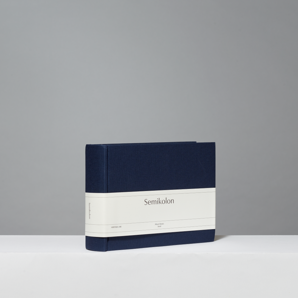 A navy blue hardcover photo album standing upright on a white surface against a light gray background. A white paper band wrapped around the middle reads 'Semikolon'.