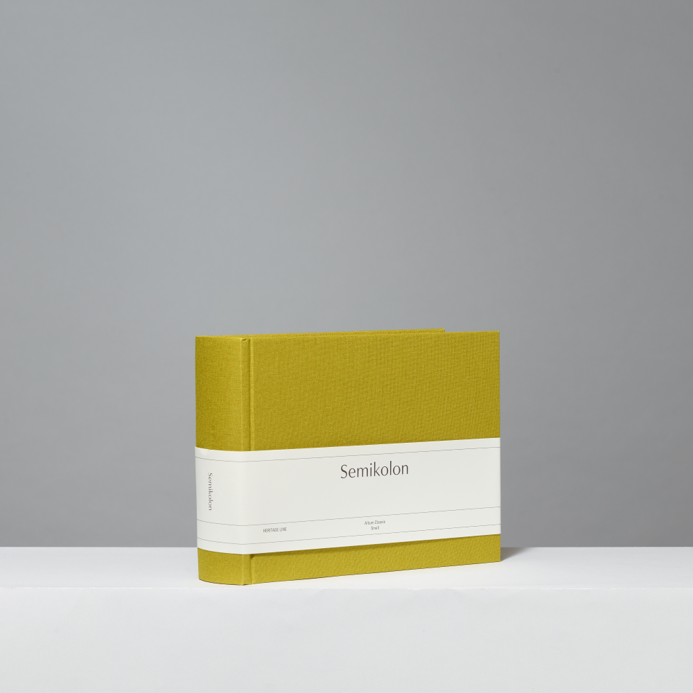 A hardcover photo album with a textured matcha cover and a white paper band around the middle, featuring the word 'Semikolon' printed in black. The book is photographed on a clean white surface against a neutral gray background.
