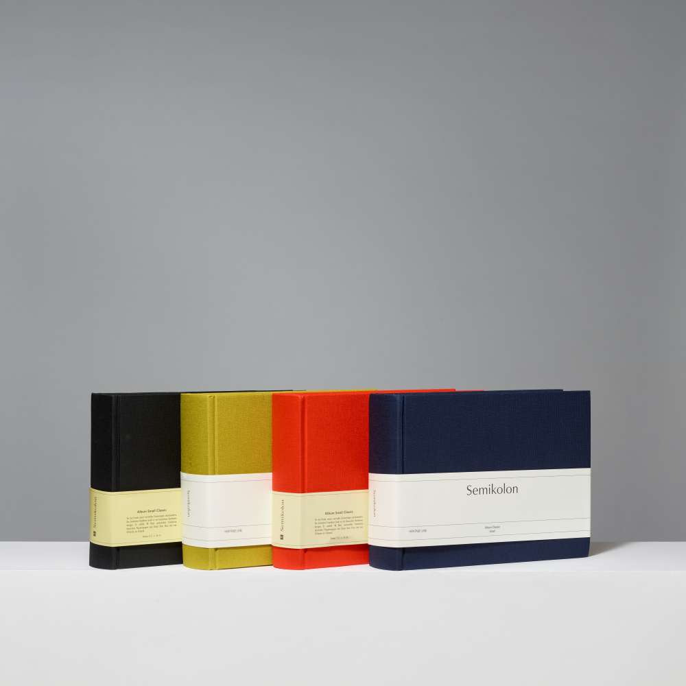 Four hardcover photo albums lined up on a white surface against a gray background. From left to right, the covers are black, matcha, orange, and blue, each with a wide white paper band around the middle.
