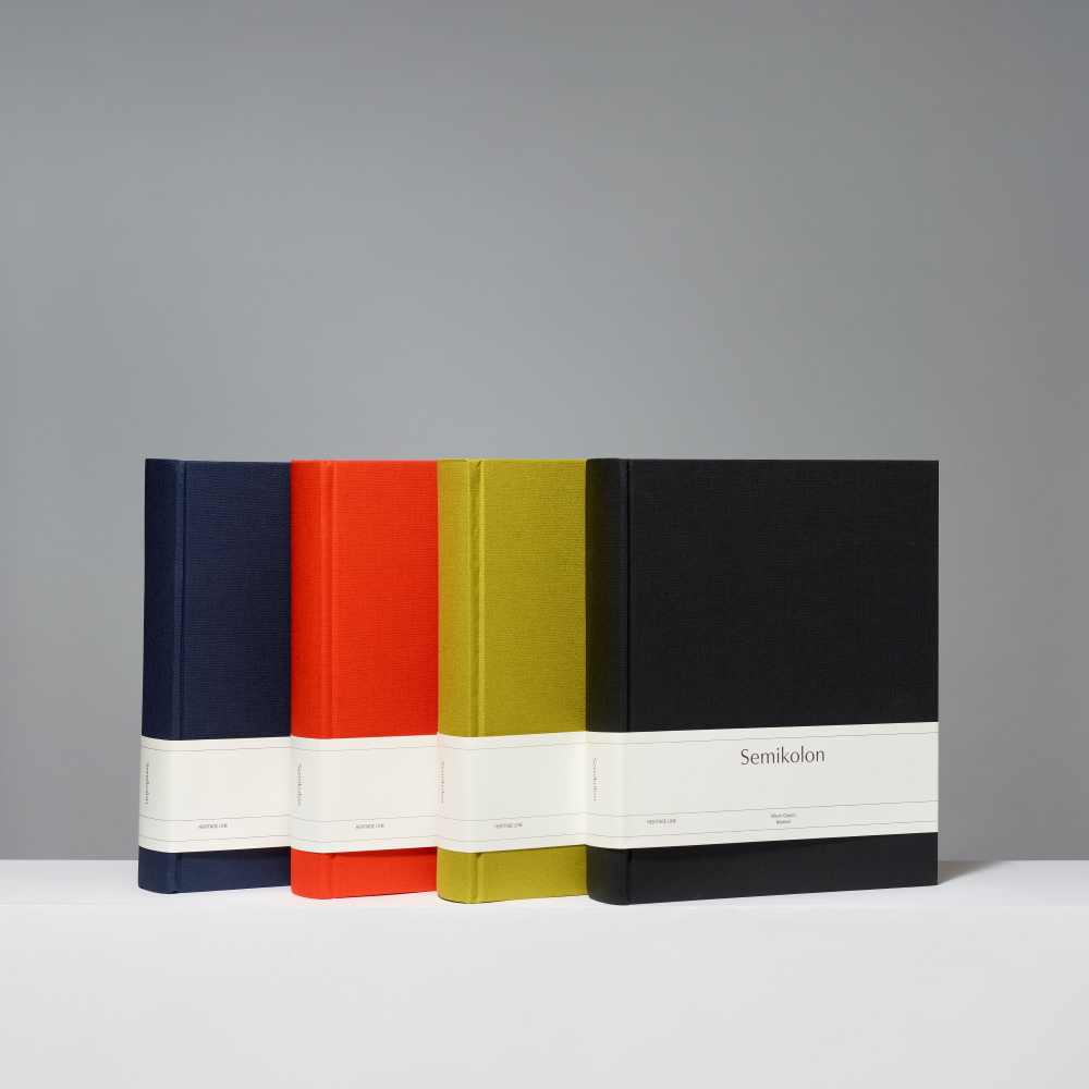 Four hardcover photo albums standing upright on a white surface against a gray studio background. The spines are in dark blue, orange, matcha, and black, and each has a wide white band around the middle with the word 'Semikolor' printed on the front band.