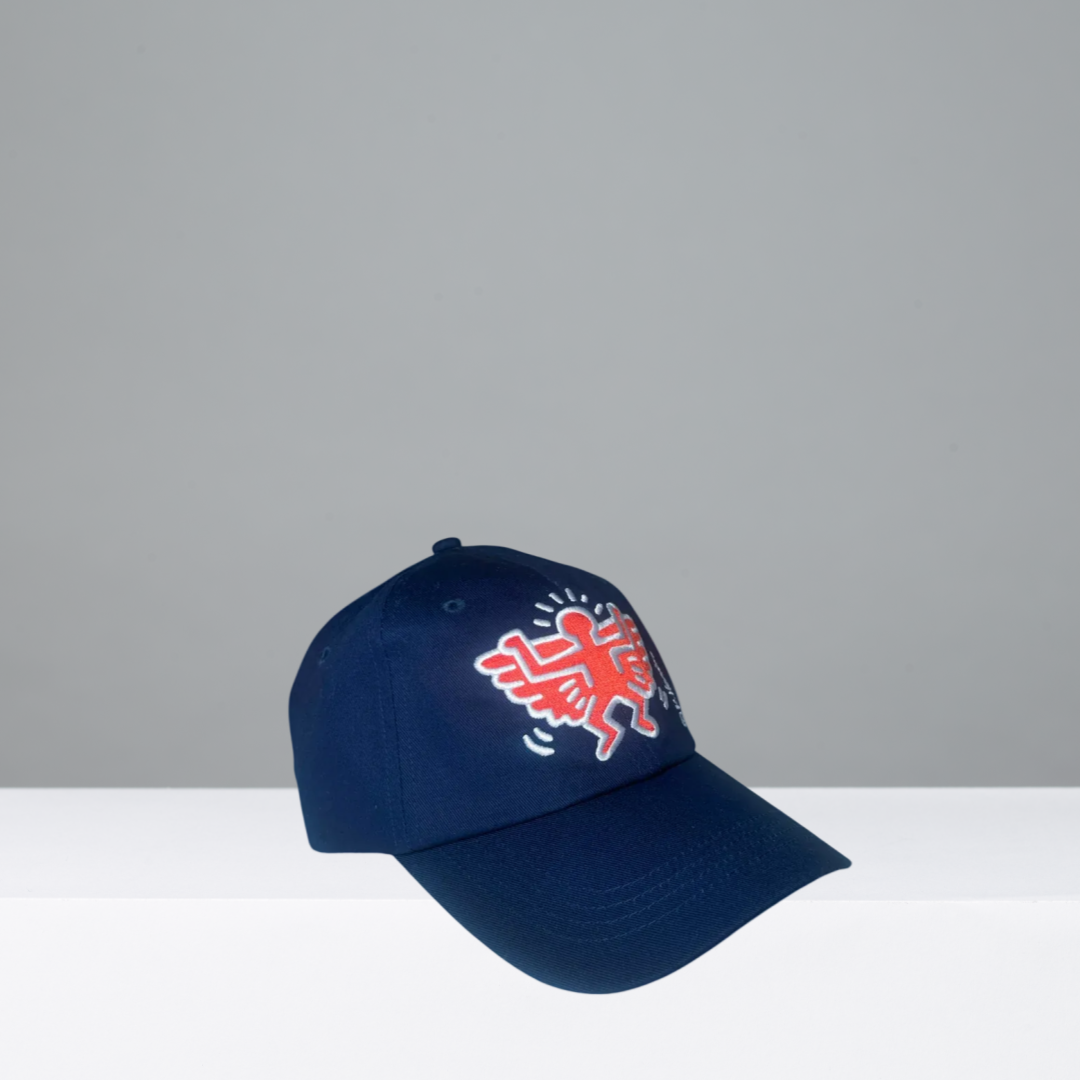 A navy blue baseball cap featuring a red and white embroidered Keith Haring style figure on the front, with a small white 'K' signature embroidered on the side, set against a gray background.