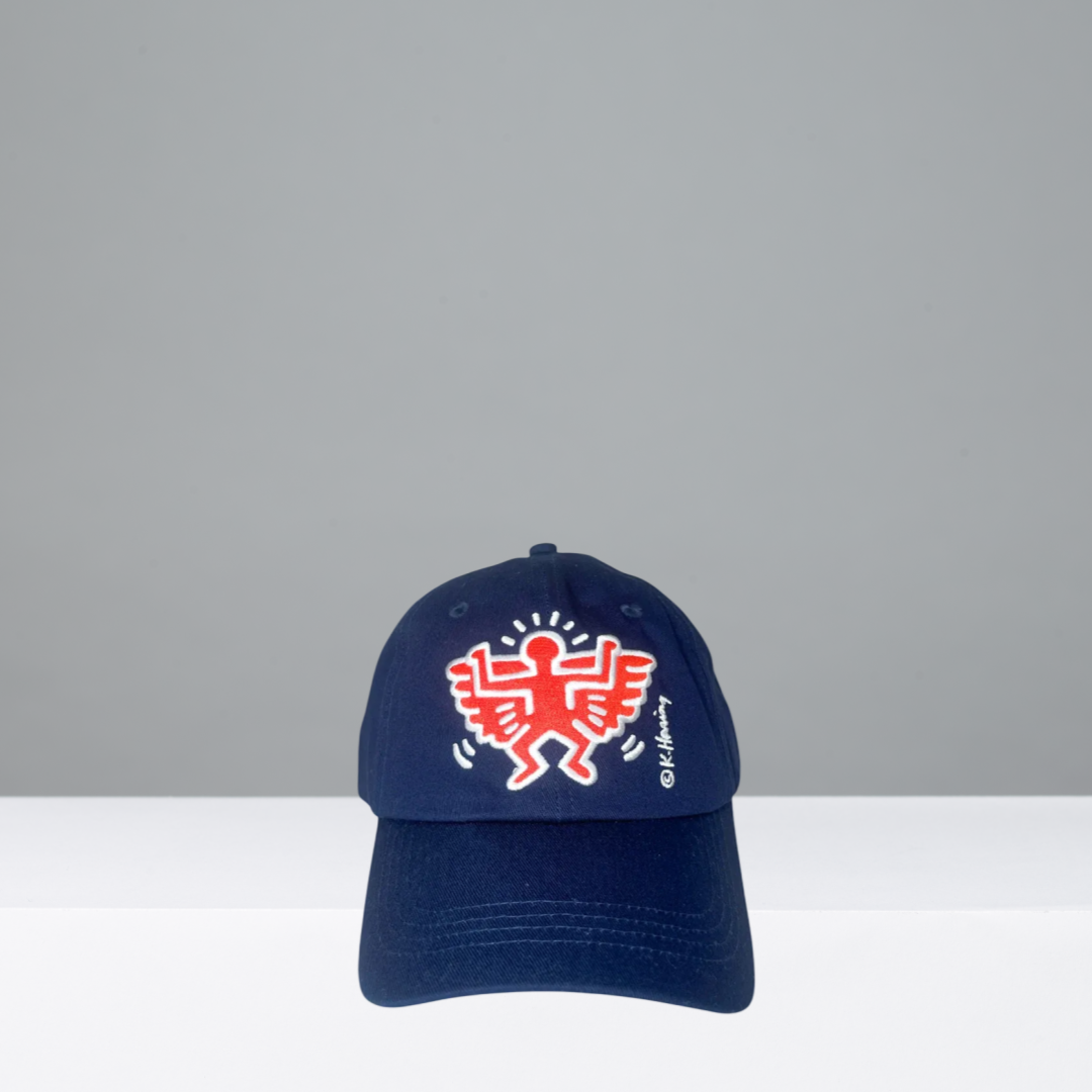 A navy blue baseball cap featuring a red and white embroidered Keith Haring style figure on the front, with a small white 'K' signature embroidered on the side, set against a gray background.