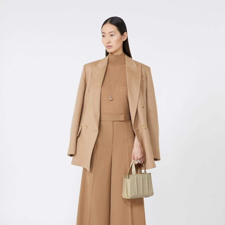 A model stands against a light gray studio background wearing a camel-colored tailored set consisting of a double-breasted blazer, matching high-neck top, and wide-leg trousers. She is holding a structured light beige handbag.