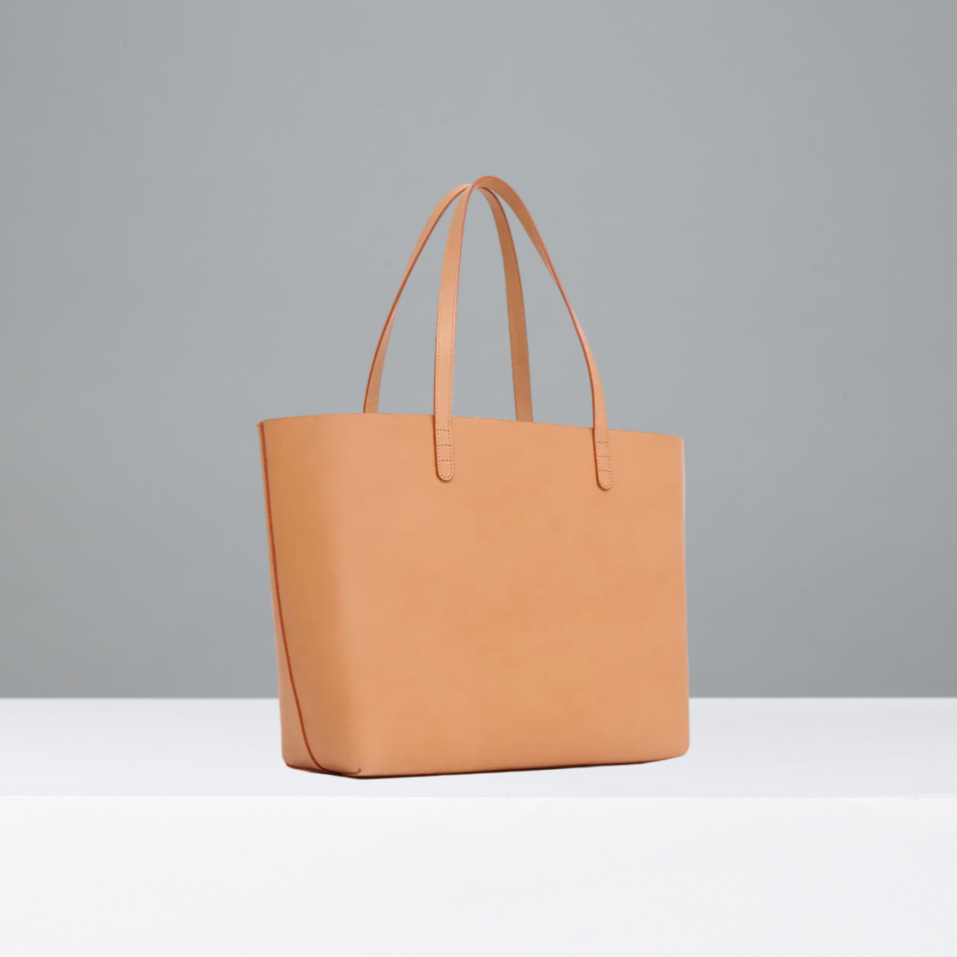 A minimalist tan leather tote bag with dual top handles, shown from a three-quarter angle against a clean, light off-white background.