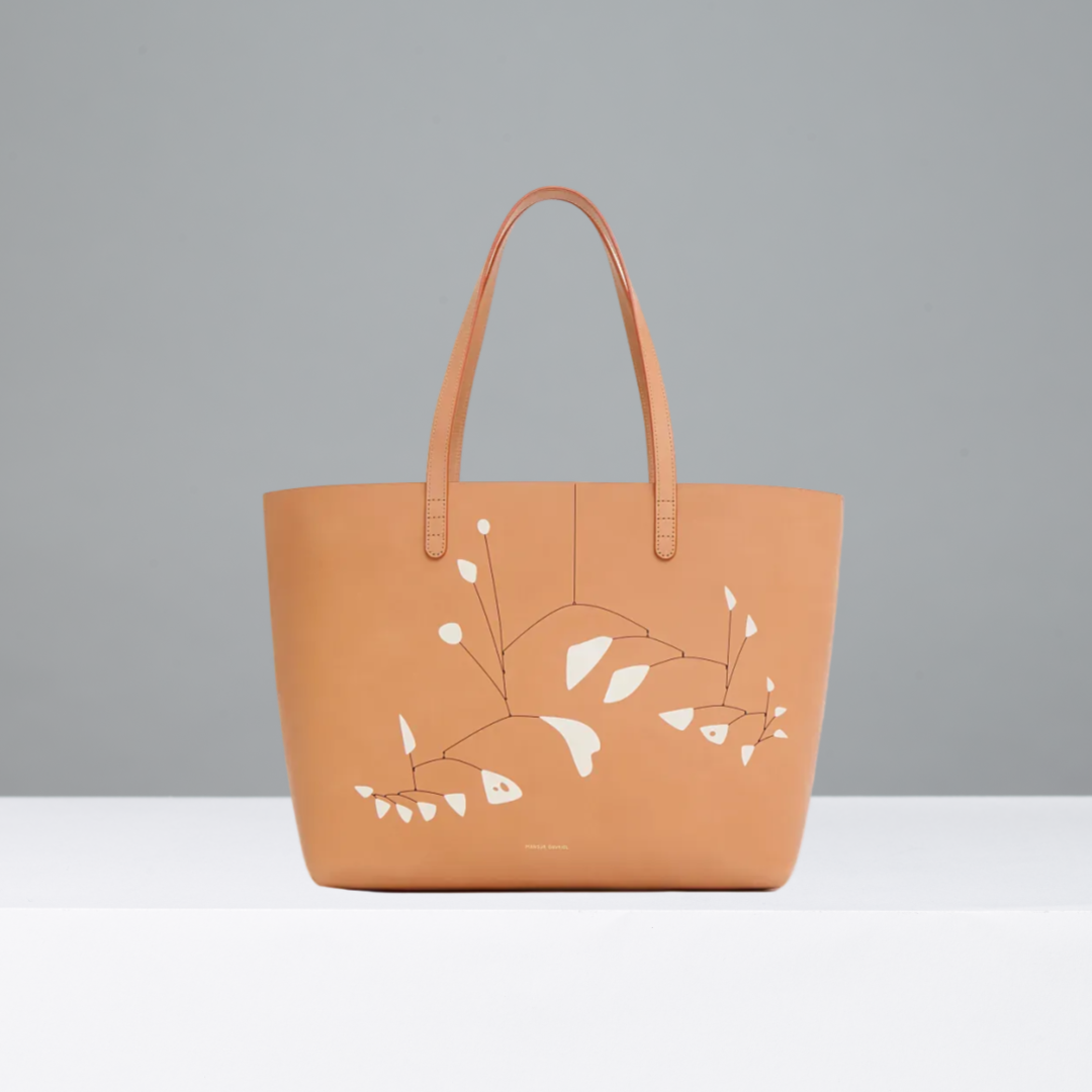 A tan leather tote bag featuring Alexander Calder's 21 feuilles blanches, with two top handles, photographed against a clean gray background.