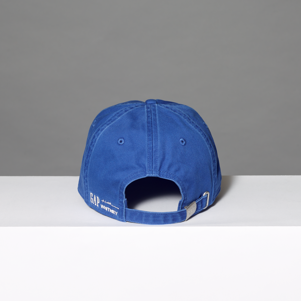 A blue cotton baseball cap shown from the back, resting on a white surface against a gray background. The back of the cap features white embroidered text reading 'GAP', 'Lichtenstein' and 'WHITNEY' alongside a silver metal buckle.