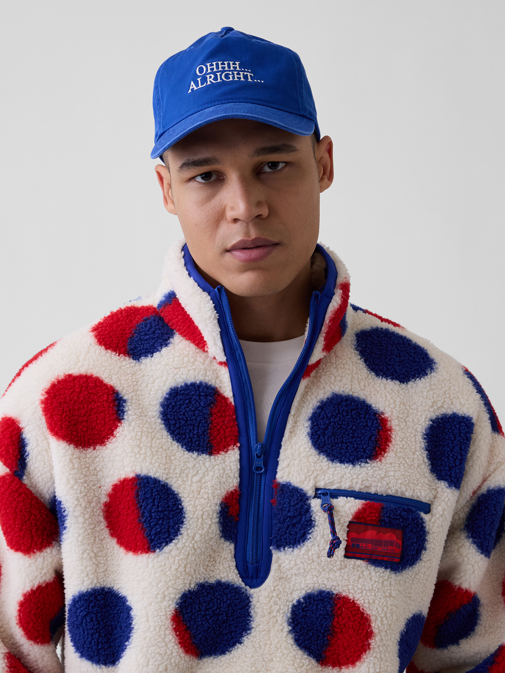 A model wearing a blue cap with Ohhh... Alright... embroidery and an off-white sherpa pullover featuring red and blue dots inspired by Roy Lichtenstein's work Cathedral #2