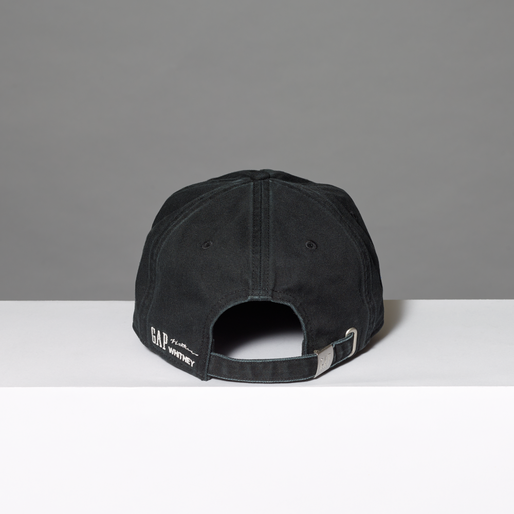 A black cotton baseball cap shown from the back, resting on a white surface against a gray background. The back of the cap features white embroidered text reading 'GAP', 'Lichtenstein' and 'WHITNEY' alongside a silver metal buckle.