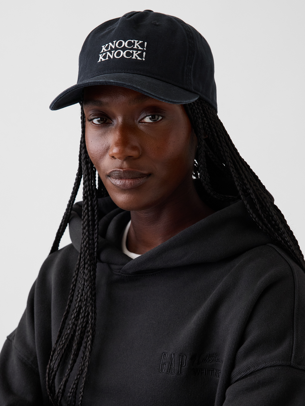 A model wearing a black baseball cap with white embroidered text reading 'KNOCK KNOCK!' inspired by Roy Lichtenstein's work Knock! Knock! Poster and a black hoodie. The photo is cropped from the chest up against a plain light gray studio background