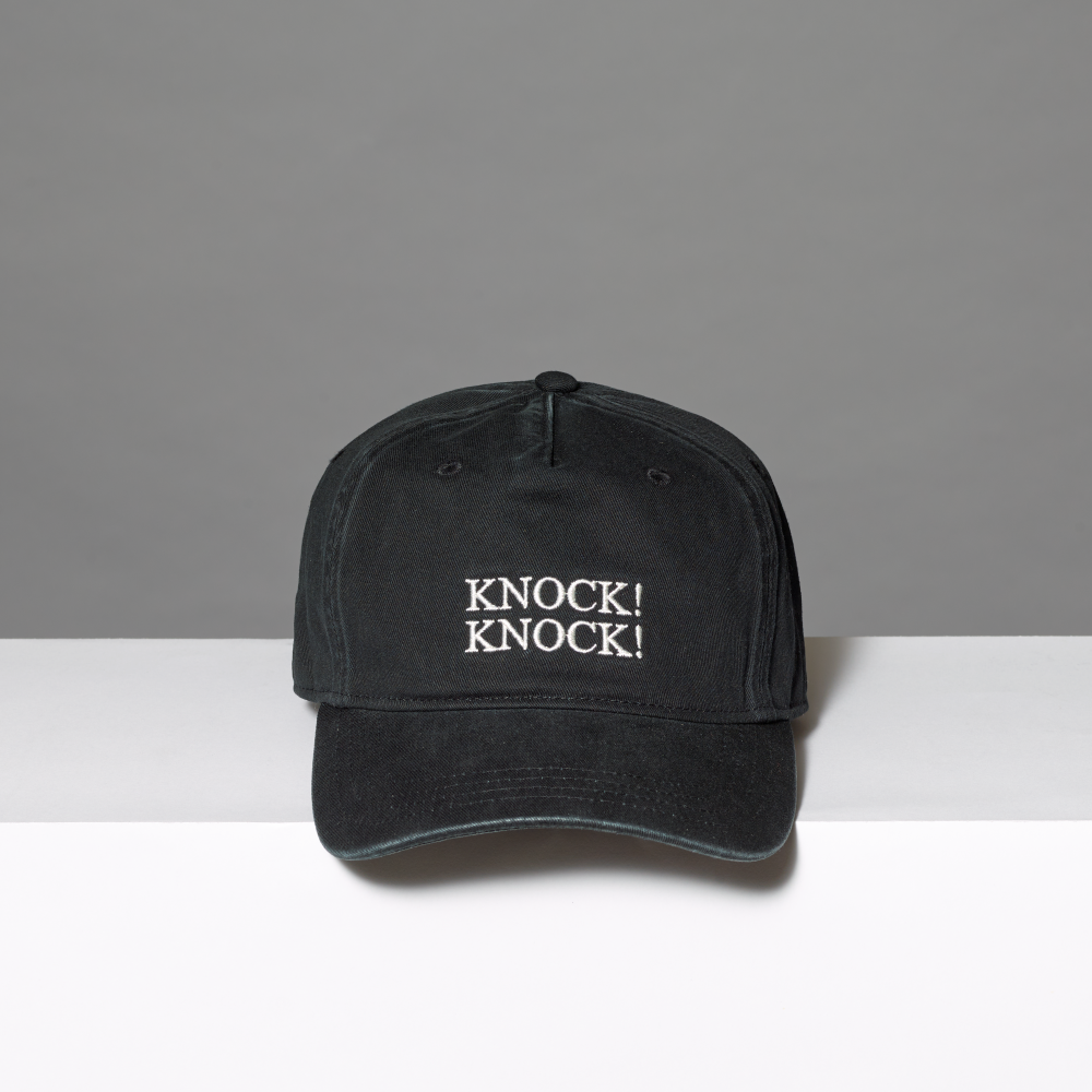 A black baseball cap with 'KNOCK! KNOCK!' inspired by Roy Lichtenstein's work of the same name, embroidered in white on the front, displayed on a white surface against a gray background