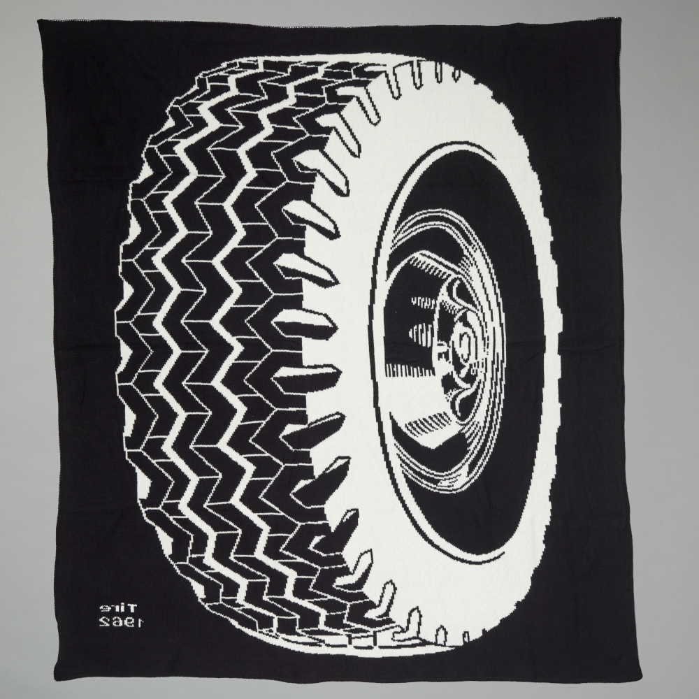 A black throw blanket featuring a bold white graphic print of a tire and wheel inspired by Roy Lichtenstein's Tire, displayed flat against a neutral gray background
