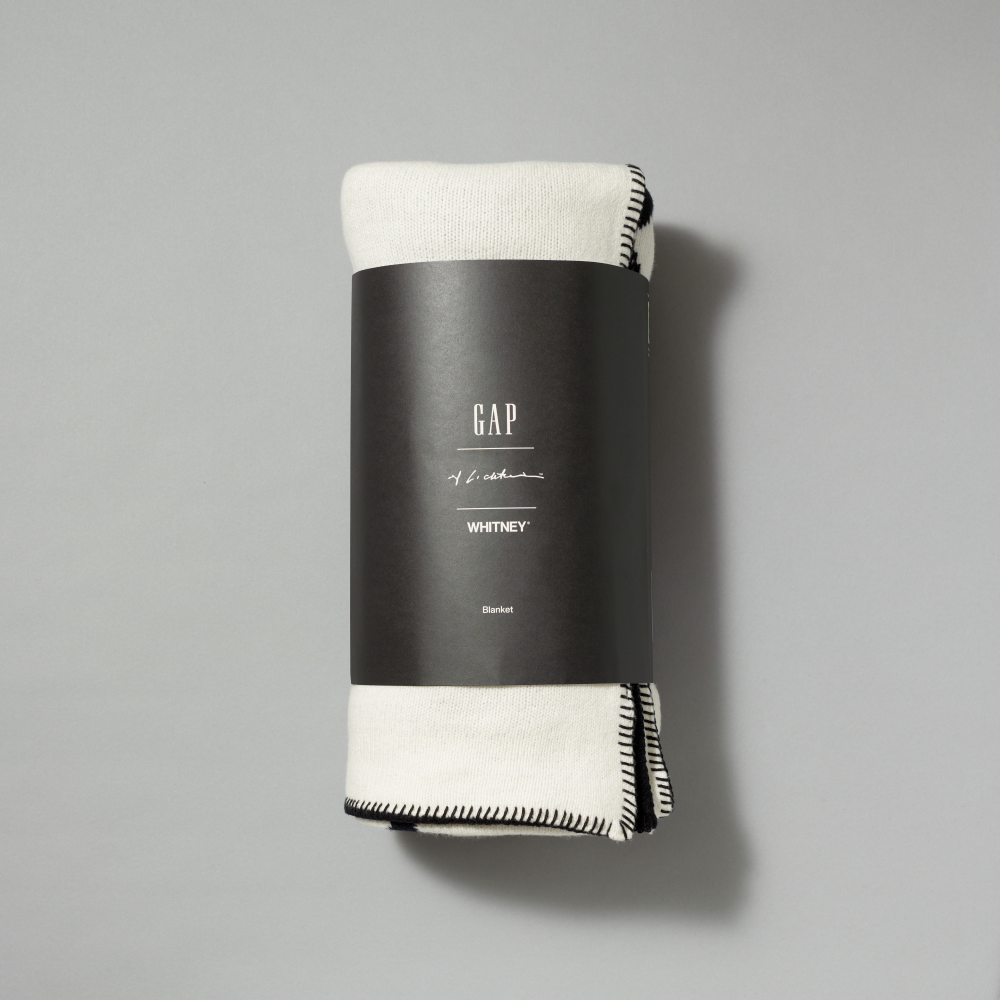 A folded off-white knit blanket with black trim, wrapped in a black paper band labeled GAP, Lichtenstein and Whitney, on a neutral gray background