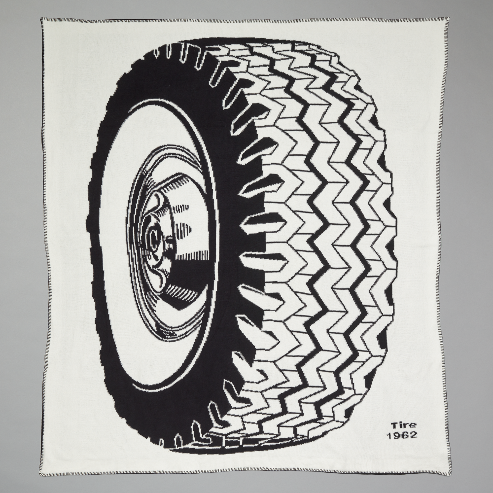 An off-white throw blanket with a large black-and-white illustration of a tire and wheel inspired by Roy Lichtenstein's work Tire shown against a light gray background