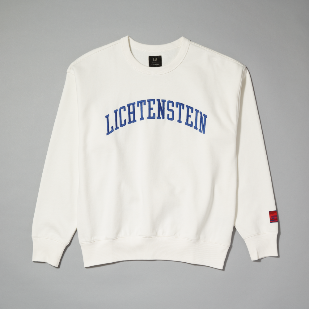 An off-white crewneck sweatshirt featuring 'LICHTENSTEIN' printed in blue arched block letters across the chest, displayed flat against a neutral gray background