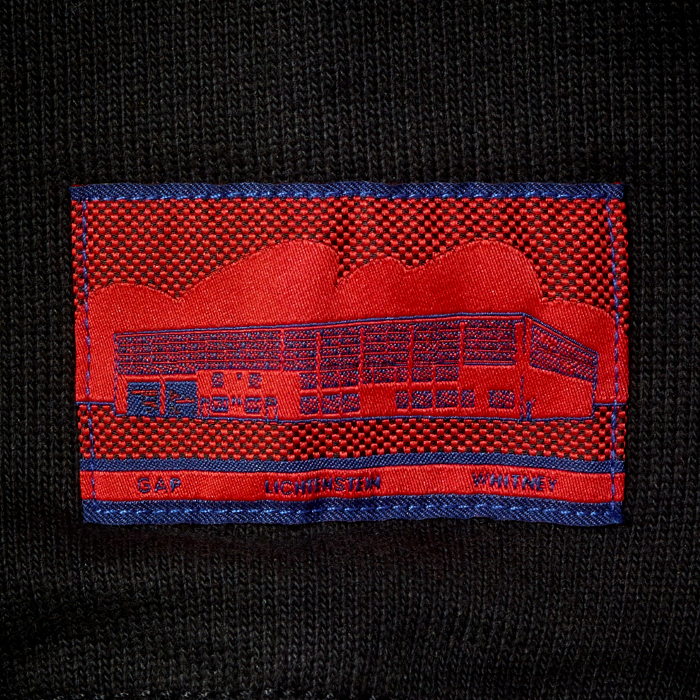Close-up of a black knit garment with a red and blue woven label featuring Roy Lichtenstein's work Real Estate and the text GAP, LICHTENSTEIN and WHITNEY