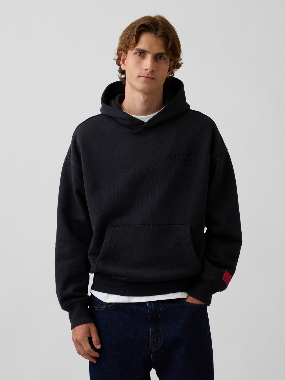A model wearing a black pullover hoodie with a tonal black logo on the left chest and a small red woven label on the cuff, paired with dark blue jeans and a white undershirt, against a light gray studio background