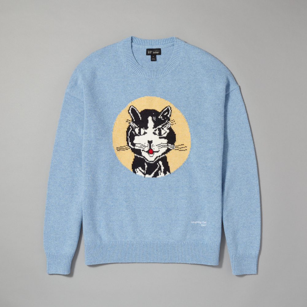 A light blue crewneck sweater featuring a large circular graphic inspired by Roy Lichtenstein's work Laughing Cat, laid flat on a neutral gray background
