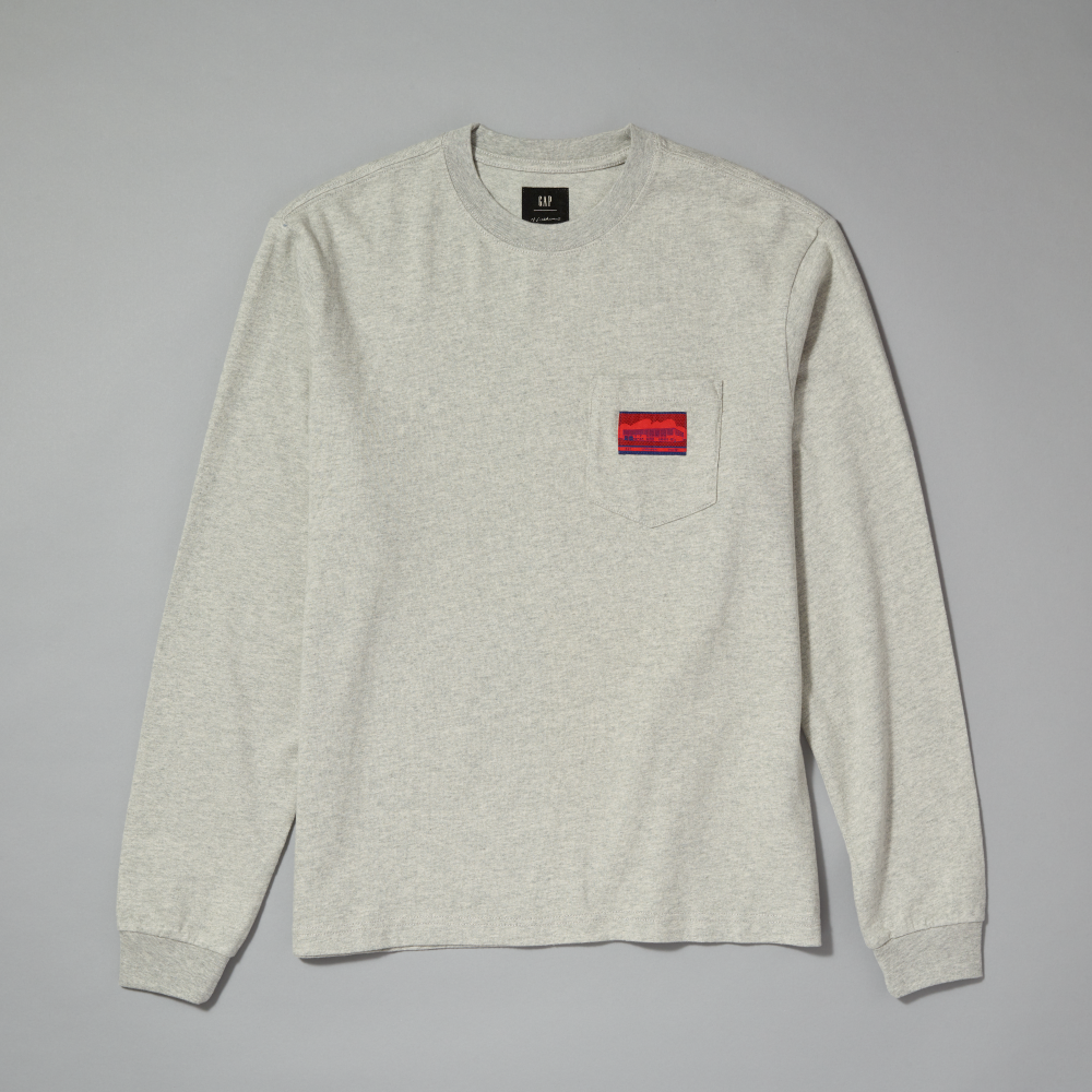 A light gray long-sleeve t-shirt with a left chest pocket featuring a red and black woven label, displayed flat against a neutral gray background