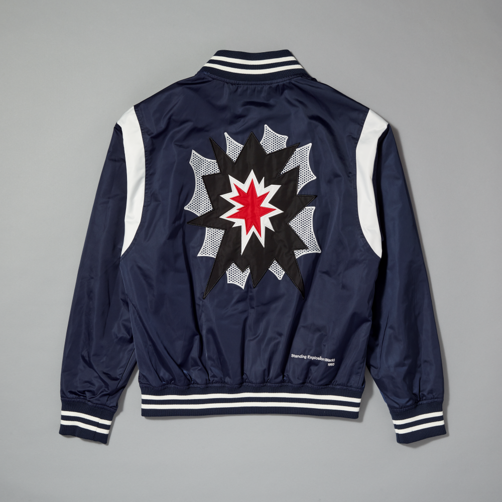 A navy blue satin varsity jacket shown from the back, featuring white shoulder panels and striped ribbed collar, cuffs, and hem. The back is decorated with a large comic-style starburst graphic inspired by Roy Lichtenstein's work Standing Explosion on a gray background
