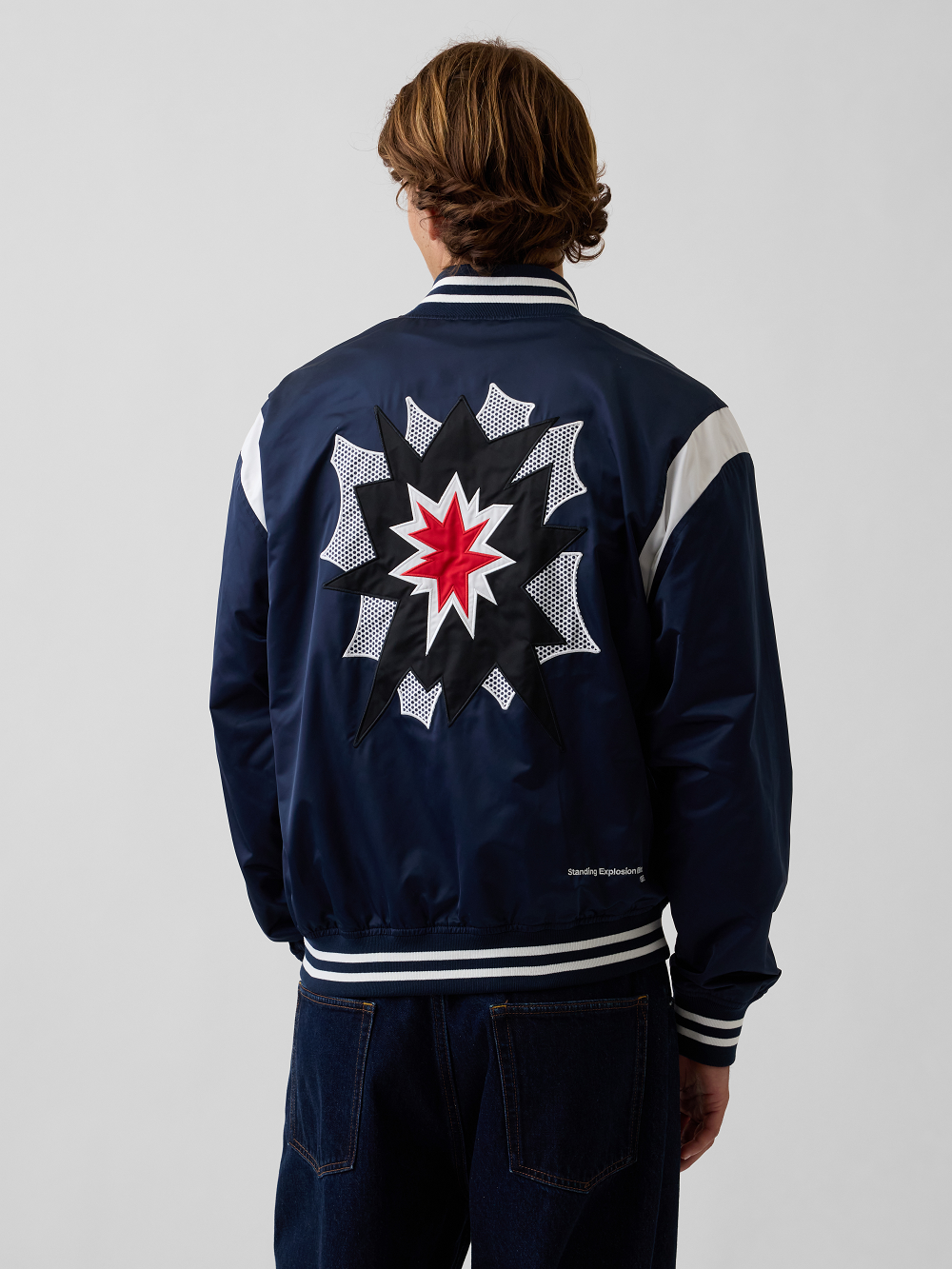 A model viewed from the back wearing a navy blue satin varsity jacket with a large graphic design inspired by Roy Lichtenstein's work Standing Explosion on the back