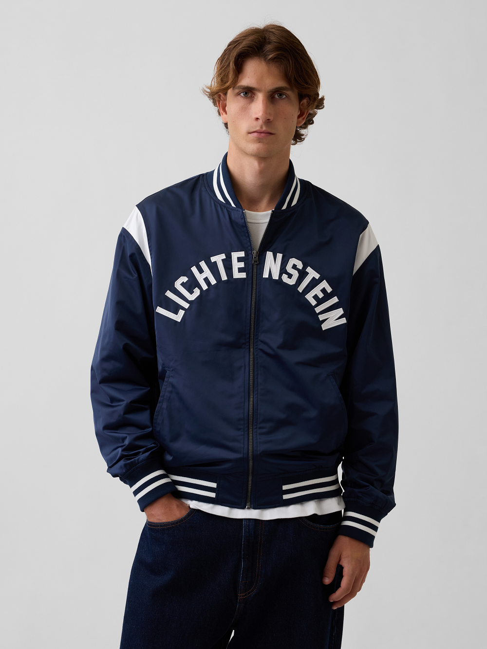 A model wearing a navy blue varsity-style varsity jacket with white stripes and 'LICHTENSTEIN' printed in white across the chest, paired with dark jeans against a light gray studio background
