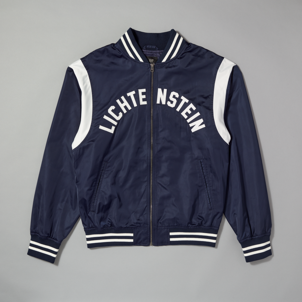 A navy blue satin varsity jacket with white stripes on the collar, cuffs, and hem, featuring white shoulder panels and the word 'LICHTENSTEIN' printed in white arched lettering across the chest on a gray background