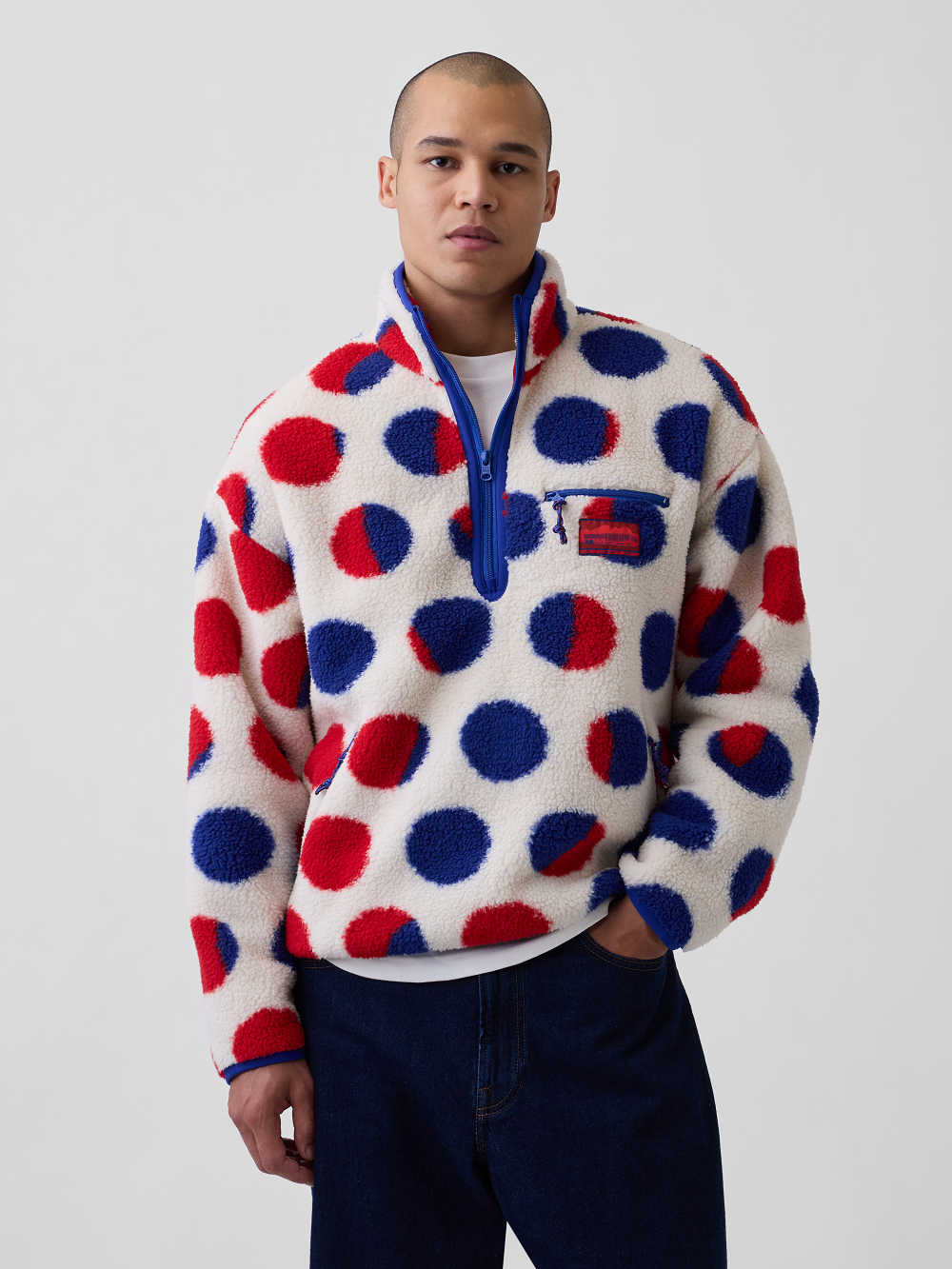A model wearing a white sherpa fleece jacket with a bold red and blue polka dot pattern inspired by Roy Lichtenstein's work Cathedral #2, paired with dark blue jeans, against a light gray studio background