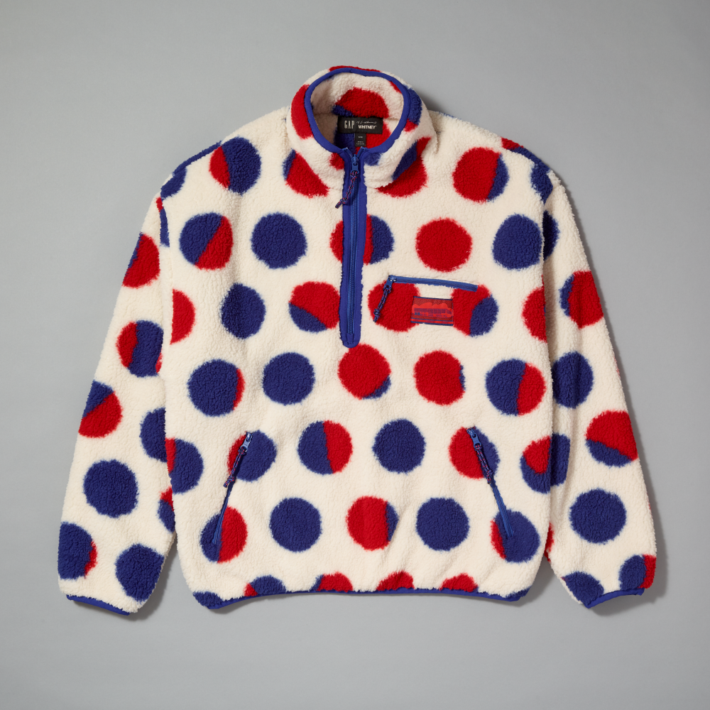 A fleece pullover sherpa with a red, white, and blue polka dot pattern inspired by Roy Lichtenstein's work Cathedral #2, featuring a blue half-zip collar, blue zipper pockets, and a red brand patch on the chest laid flat against a gray background