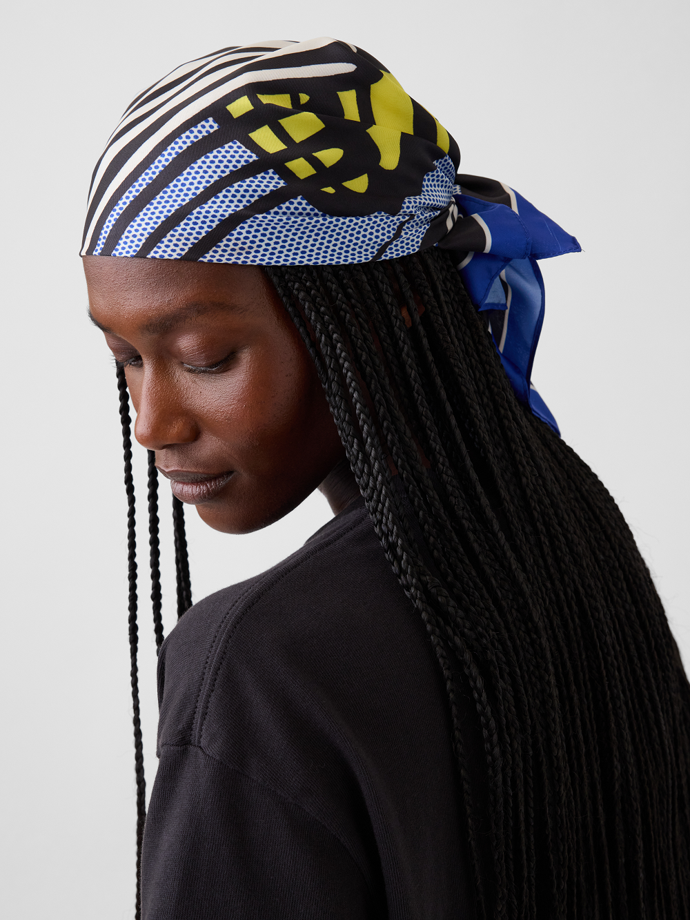 A model shown in side profile wearing a patterned headscarf inspired by Roy Lichtenstein's work Little Big Painting wearing a black top against a light gray studio background