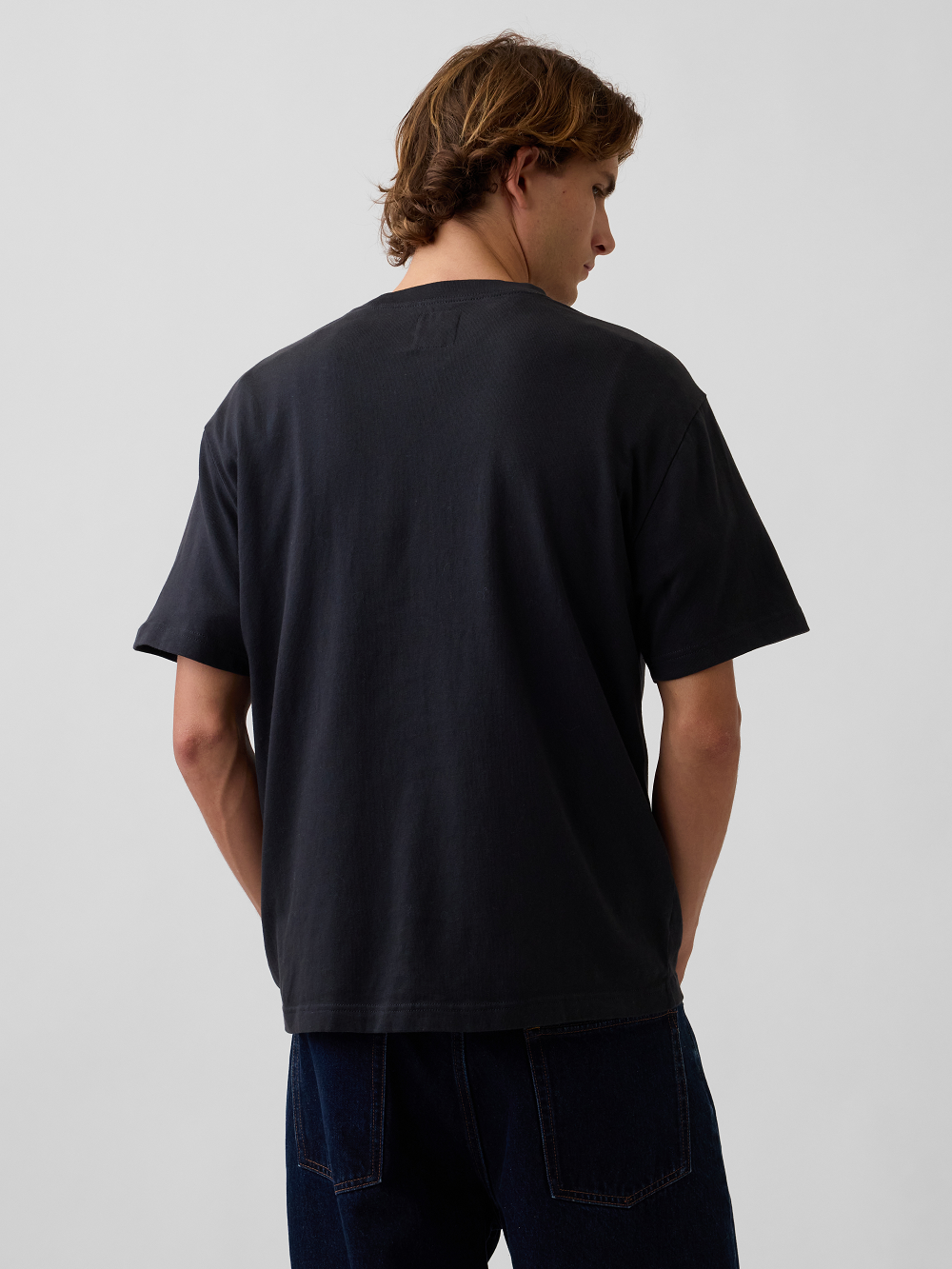 A model viewed from behind wearing a black short-sleeve t-shirt and dark blue jeans, standing against a light gray studio background