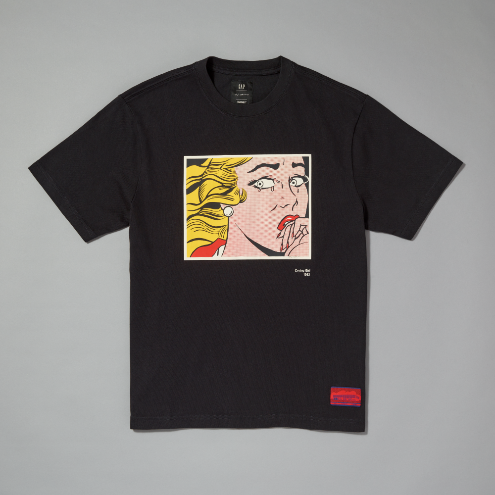 A black short-sleeve crewneck t-shirt featuring a graphic print inspired by Roy Lichtenstein's work Crying Girl on the chest, with a red and blue woven label on the lower right hem, shown flat against a neutral gray background
