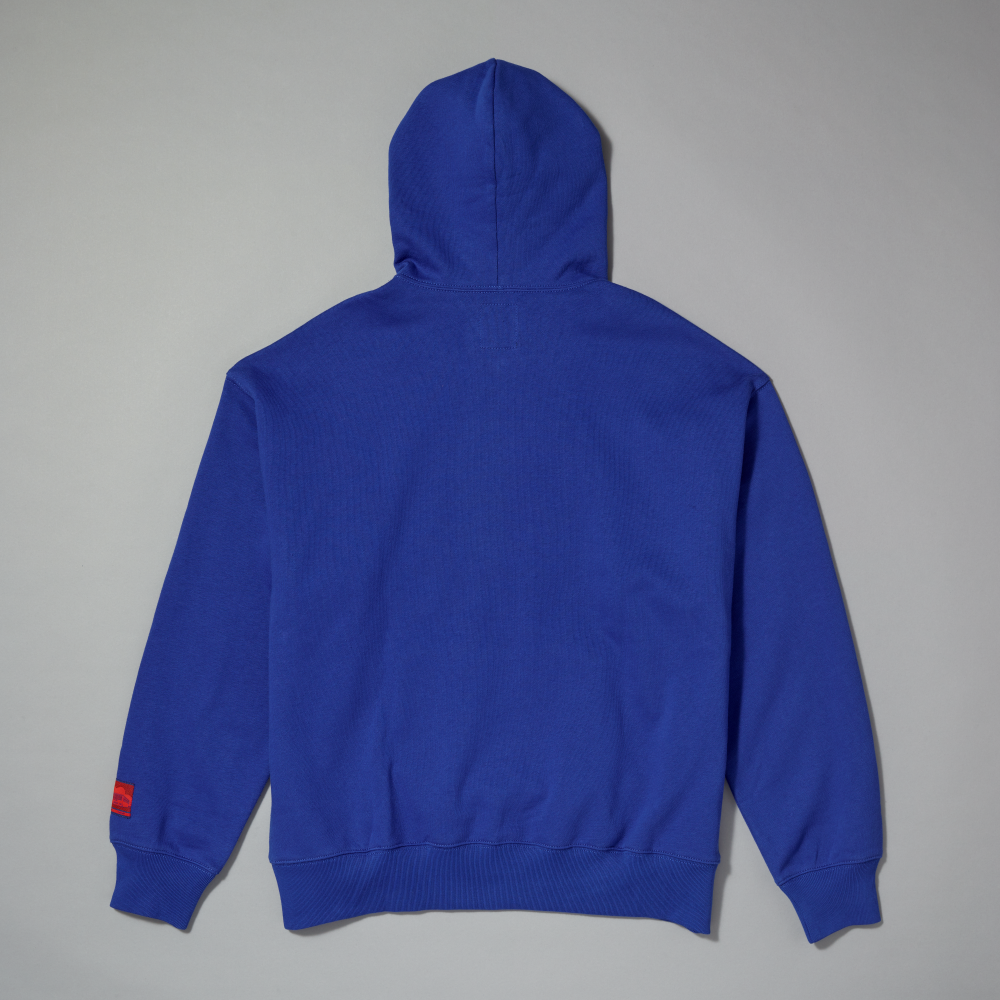 The back view of a royal blue hooded sweatshirt with a ribbed hem and cuffs, featuring a small red woven label on the left sleeve, laid flat against a neutral gray background
