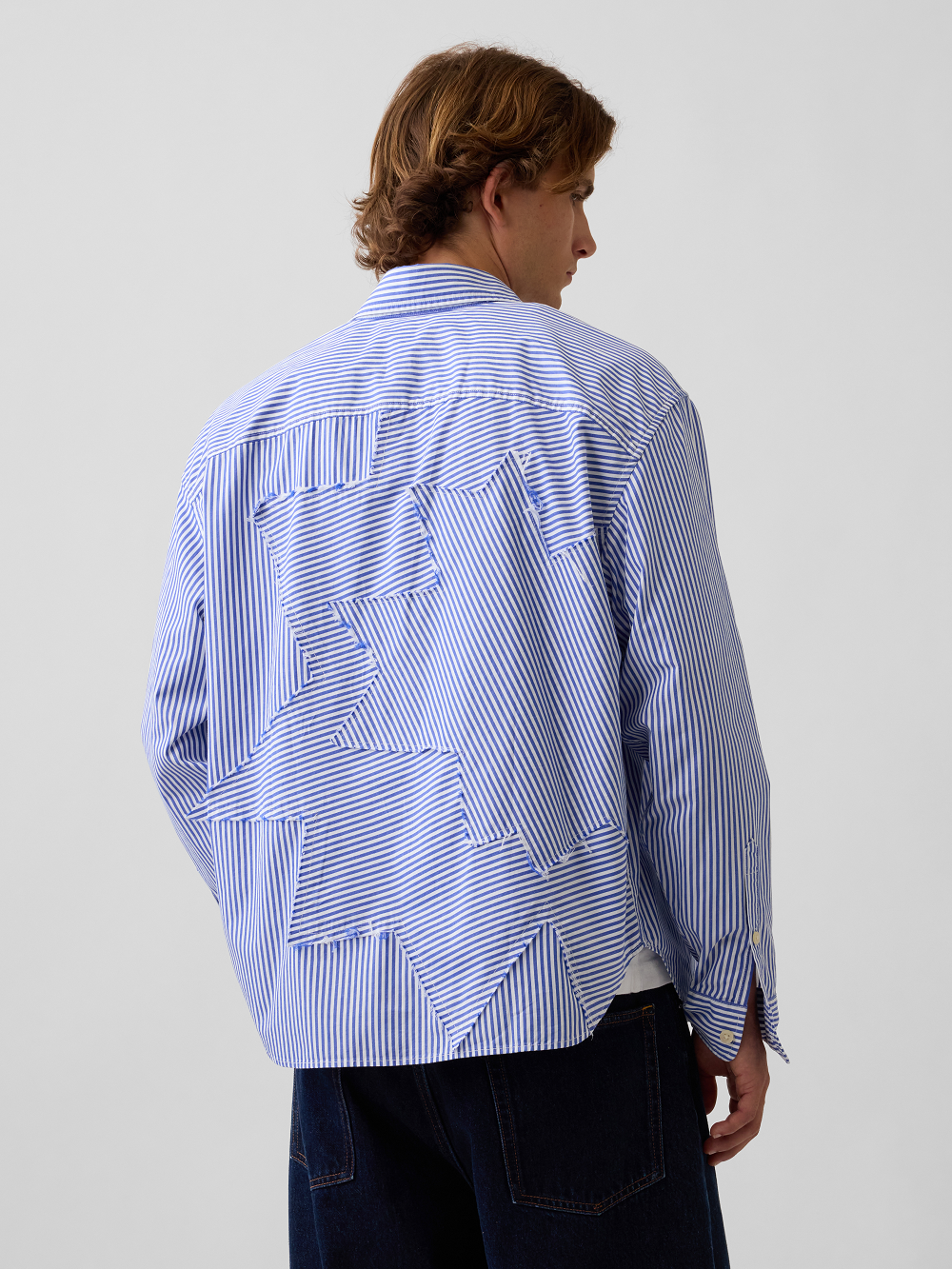 A model viewed from the back wearing a light blue and white striped button-up shirt with a geometric patchwork pattern inspired by Roy Lichtenstein's work Standing Explosion on the back, paired with dark blue jeans, against a plain light gray studio background