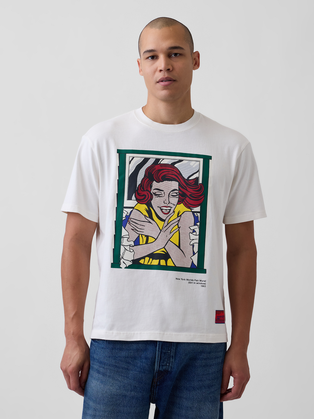 A model wearing a white graphic t-shirt inspired by Roy Lichtenstein's work Girl in Window, paired with blue jeans, against a light gray background